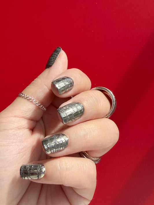 Hand with silver croc nail polish and rings against a red background